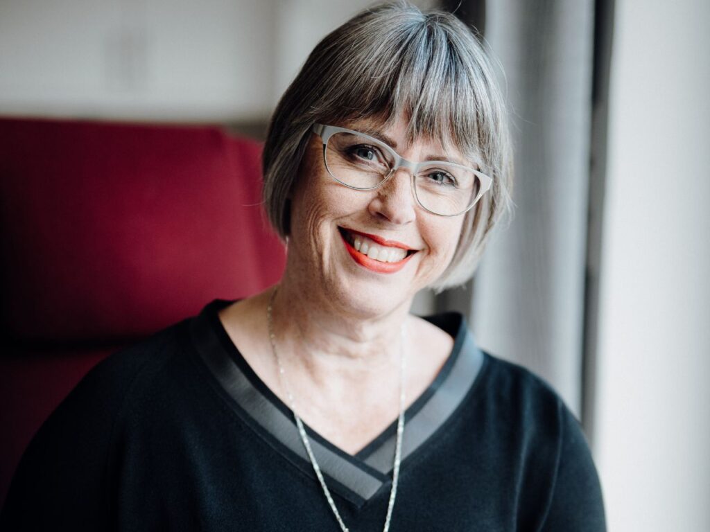 Karin Stehr lächelnd mit silberner Brille im roten Sessel mit 